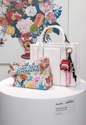 Two bags displayed on a pedestal: a colorful floral handbag with "Never Stop Growing" text and a white structured tote with a heart-shaped keychain.