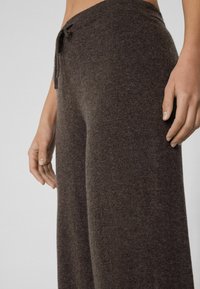 Brązowe dzianinowe spodnie typu wide-leg o miękkiej fakturze, z elastycznym pasem i luźnym fasonem. Zbliżenie na pas i rękę dla szczegółów.