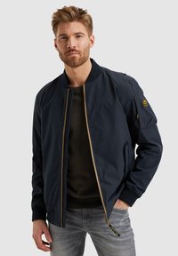 Chaqueta bomber navy con cierre dorado, cuello acanalado y bolsillos laterales. Presenta un parche de emblema en la manga izquierda y una textura suave.