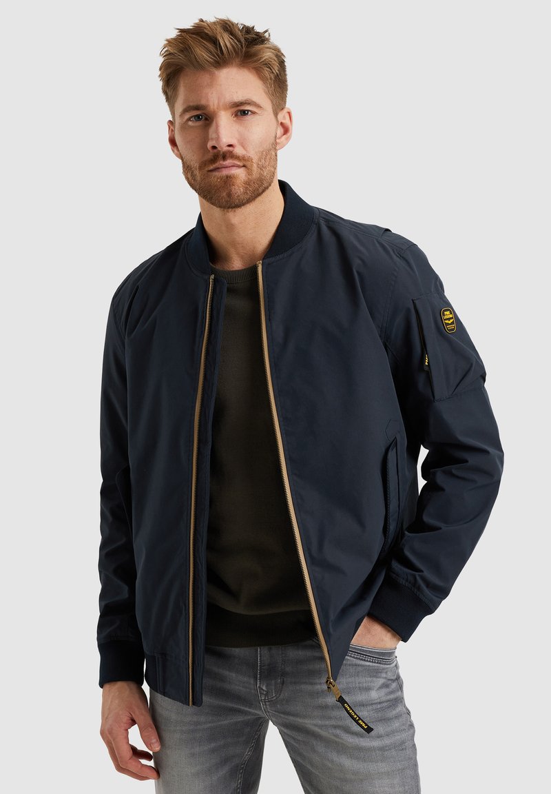Chaqueta bomber navy con cierre dorado, cuello acanalado y bolsillos laterales. Presenta un parche de emblema en la manga izquierda y una textura suave.