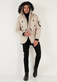 Jachetă umplută de culoare bej deschis, cu glugă căptușită cu blană, accente negre, buzunare cu fermoar și margine cu șnur; purtată cu pantaloni negri slim-fit și cizme.