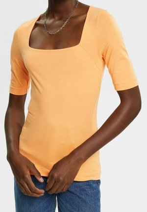 Person trägt ein eng anliegendes pfirsichfarbenes kurzärmliges Oberteil mit quadratischem Ausschnitt und blaue Jeans, Hände nahe der Taille positioniert.