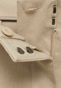 Tissu beige avec un bouton, une fermeture éclair et deux clips en métal foncé. Gros plan montrant les détails de couture et de texture.