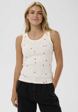 Lyshåret kvinde iført hvid ærmeløs tanktop med små røde hjerte-mønstre og sorte bukser, stående med den ene hånd i lommen.