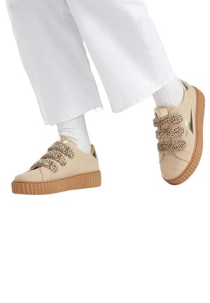 Beige sneakers met drie Velcro-straps met luipaardprint, metallic gouden accenten en een textuur gumzool, gecombineerd met witte wijde broek.