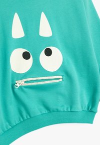 Felpa turchese con una faccia da cartone animato bianca, caratterizzata da grandi occhi e due orecchie appuntite. Include una piccola tasca con zip sul davanti.