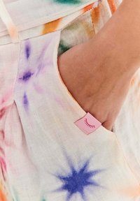 Ruka odpočívajúca v vrecku svetlých nohavíc s farebnými vzormi tie-dye hviezd a malou ružovou nálepkou s návrhom zatvoreného oka.