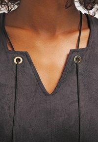 Schwarzes Wildledershirt mit V-Ausschnitt und goldenen Ösen. Verfügt über dünne schwarze Kordeln zur Anpassung entlang des Ausschnitts.
