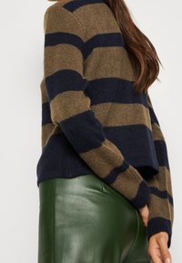 Hnědo-modrý pruhovaný svetr s pletenou strukturou, má spadlá ramena a zkrácený střih, nošený přes zelené kožené kalhoty.