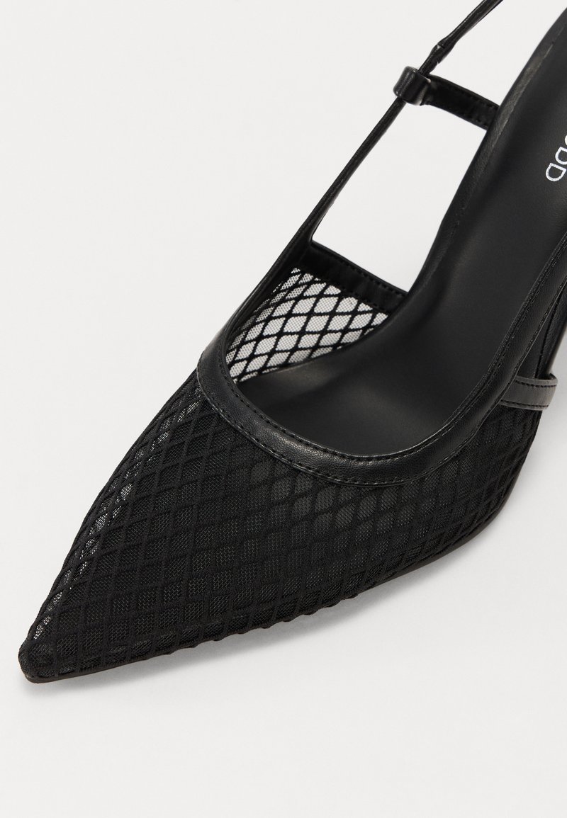 Chaussure à talons hauts noire avec un bout pointu, présentant un design en maille et des accents en cuir. Le talon est fin et la chaussure a un panneau latéral transparent.