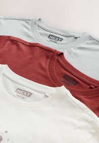Trois t-shirts en coton gris, bordeaux et blanc, avec des encolures rondes et des étiquettes de marque visibles, empilés proprement.