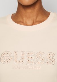 Camiseta de manga corta de color rosa claro con la inscripción "GUESS LOS ANGELES" en letras en relieve y punteadas, adornada con pequeños studs plateados.