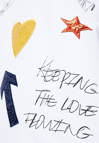 Patchs à paillettes en forme de cœur jaune, étoile rouge et flèche bleue avec texte manuscrit « KEEPING THE LOVE FLOWING » sur tissu blanc.
