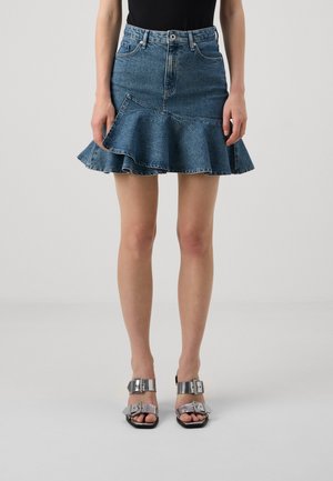 Mujer con falda vaquera azul con dobladillo con volantes y sandalias de tacón alto con hebilla plateada, de pie frente a un fondo gris liso.