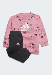 Felpa rosa con motivi a doodle neri e logo Adidas bianco, abbinata a pantaloni della tuta neri con piccolo logo rosa sulla gamba.