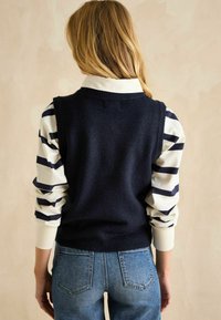 Marineblauer Weste aus Strickstoff über einem gestreiften Langarmshirt. Dunkelblaue Jeans, sichtbare Nähte und Gesäßtaschen. Der Gesamteindruck ist glatt und angenehm.