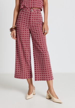 Femme portant un pantalon large court bordeaux et un haut assorti avec un motif chaîne blanche, accessoirisée de bagues, bracelets et talons blocs beiges.