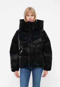 Černá péřová bunda s lesklým povrchem, oversized límcem a prošívaným designem. Model nosí modré džíny, čímž ukazuje neformální styl.