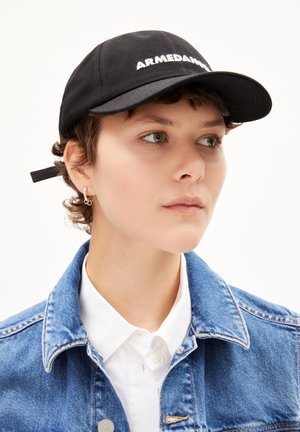 Cappellino nero con "ARMEDANGELS" in bianco, realizzato in tessuto, dalla forma classica. Indossato con una giacca di jeans blu e una camicia bianca.