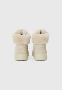 Bottes d'hiver beige avec des bordures duveteuses, pr�ésentant une finition texturée et un logo au talon, sur un fond blanc uni.