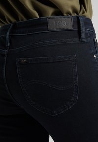 Jeans de un azul oscuro, confeccionados en denim, con una etiqueta de cuero en la cinturilla y un bolsillo que presenta un detalle de costura curva.