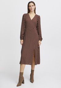 Robe brune à pois comportant des manches longues, un décolleté en V et une fente à l'avant. Associée à des bottes à talons hauts beige. Texture de tissu lisse.