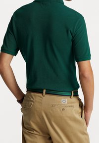 Mörkgrön pikétröja med korta ärmar, i kombination med beige byxor som har en flerfärgad rand i midjan och detaljer med bakfickor.