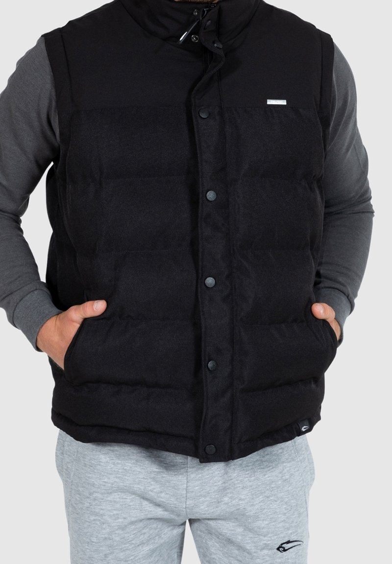 Homme portant un gilet matelassé noir avec des boutons-pression sur une chemise grise à manches longues, les mains dans les poches du gilet, assorti à un pantalon de survêtement gris clair.