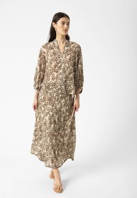 Langes, fließendes Kleid in hellbeige mit einem braunen abstrakten Muster, ausgestattet mit einem V-Ausschnitt und Puffärmeln. Das leichte Gewebe fällt geschmeidig.