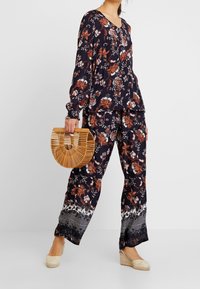 Frau trägt eine marineblaue Bluse mit Blumenmuster und passende Hose, hält eine Handtasche aus Holzlatten und trägt beigen Keilabsatz-Espadrilles.