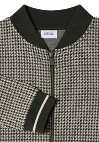 Veste bomber avec motif pied-de-poule noir et blanc, col et poignets côtelés noirs avec une bande blanche, et fermeture éclair à l'avant.