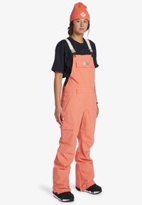 Heldere koraalkleurige tuinbroek met verstelbare witte banden, zijzakken en een frontzak, gecombineerd met een zwart T-shirt en een oranje muts.