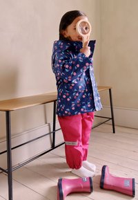 Zils lietusmētelis ar rozā ķiršu rakstu, kopā ar rozā ūdensnecaurlaidīgām biksēm. Bērns tur palielināmo stiklu, uz grīdas ir rozā lietus zābaki.