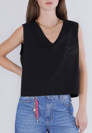 Donna che indossa un top nero senza maniche con scollo a V e logo discreto, collane d'oro a strati, jeans blu e un portachiavi rosa con ciondolo.