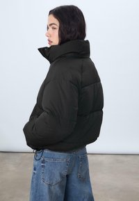 Giubbotto imbottito nero con colletto alto, caratterizzato da un design trapuntato e cordino elastico in vita, abbinato a jeans azzurri chiari.