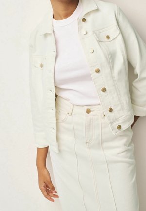 Personne portant une veste en denim blanche à boutons dorés, un t-shirt blanc cintré et une jupe blanche taille haute avec des coutures à l'avant.