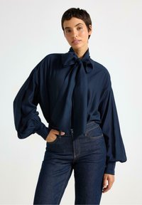 Marineblå bluse med overdimensionerede ærmer og en stor sløjfe ved kraven, parret med mørke denimjeans. Glat stof med en subtil glans.