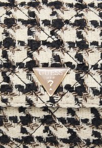 Driehoekig metalen Guess U.S.A. logo op gestructureerde zwart, bruin en crème geweven stof met een geometrisch patroon.