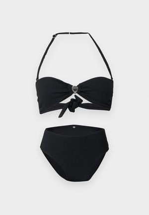 Woman standing barefoot in a black two-piece swimsuit with high-waisted bottoms, looking slightly to the side against a plain background.