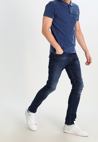 Marineblaues Baumwoll-Poloshirt mit Kragen und Tasche, kombiniert mit dunkelblauen Slim-Fit-Jeans und weißen Sneakern. Glatte Textur und klassisches Design.