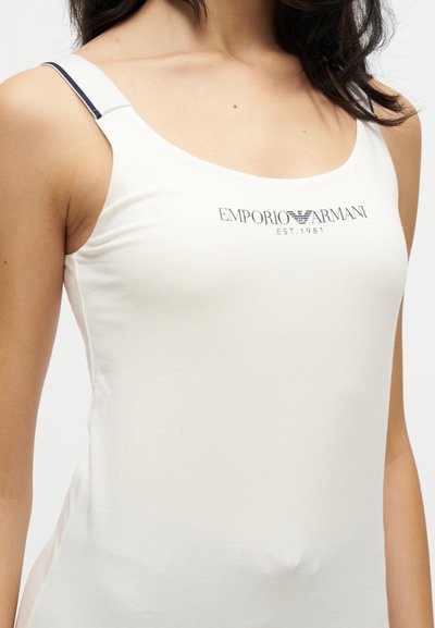 Γυναίκα με λευκό αμάνικο τοπ με το λογότυπο Emporio Armani και ρυθμιζόμενους μαύρους ριγωτούς ιμάντες σε μονόχρωμο φόντο.