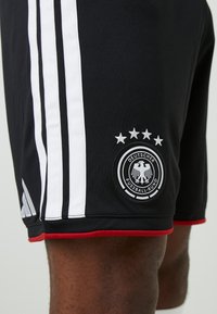 Čierne športové šortky s bielymi vertikálnymi pruhmi, červenými akcentmi a vyšitým kruhovým nášivkom s logom Nemeckej futbalovej asociácie.