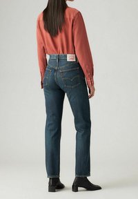 Světle rezavá košile s dlouhými rukávy, zkombinovaná s tmavými džínami s vysokým pasem. Černé kotníkové boty dotvářejí celý outfit.