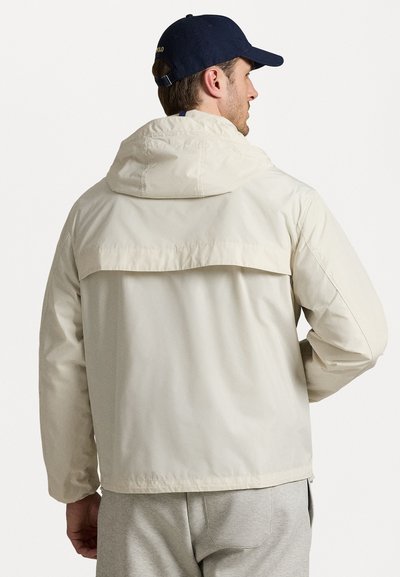 Jachetă cu glugă de culoare bej deschis, cu o croială lejeră, fabricată din material impermeabil. Dispune de o spate cu yoke și manșete elastice. Asortată cu pantaloni gri.