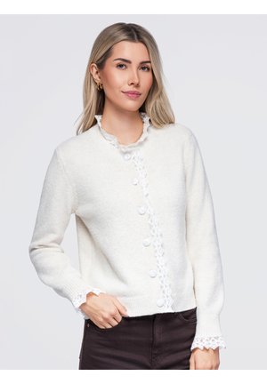 Une femme porte un cardigan blanc boutonné sur le devant, orné d’une bordure en dentelle, avec un pantalon foncé, sur un fond clair.