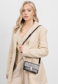 Borsa a tracolla nera con texture, con una chiusura in argento, forma rettangolare e tracolla regolabile, indossata sopra un giubbotto beige.