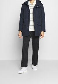 Mörkblå vattentät jacka med huva, med dragkedja och knäppning, två framfickor, bärs över en randig polo och svarta byxor.