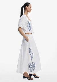 Vit cropped topp med puffärmar som har blå bladbroderi; lång vit kjol med matchande blå spiral mönster; svarta klacksandaler.