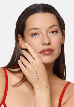 Woman with brown hair and light skin wearing a red strap top, resting her hand with a silver chain bracelet featuring small crystals on her face.
