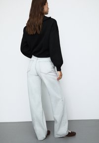 Črna prevelika sekcija, svetlo siva široka jeans hlače, sproščeni rjavi čevlji. Vidni zadnji žepi na hlačah, teksturirana tkanina puloverja in subtilni kovinski dodatki.